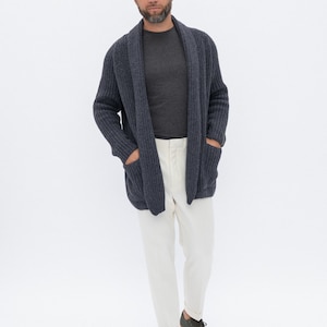 May include: A man wearing a dark blue cardigan over a gray t-shirt, white pants, and dark green sneakers.