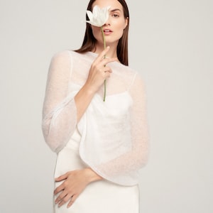 May include: A white sheer knit bolero with long sleeves and a wide neckline, worn over a solid white sleeveless top. The garment features a lightweight, semi-transparent texture and a clean, simple design that sits comfortably over the shoulders.