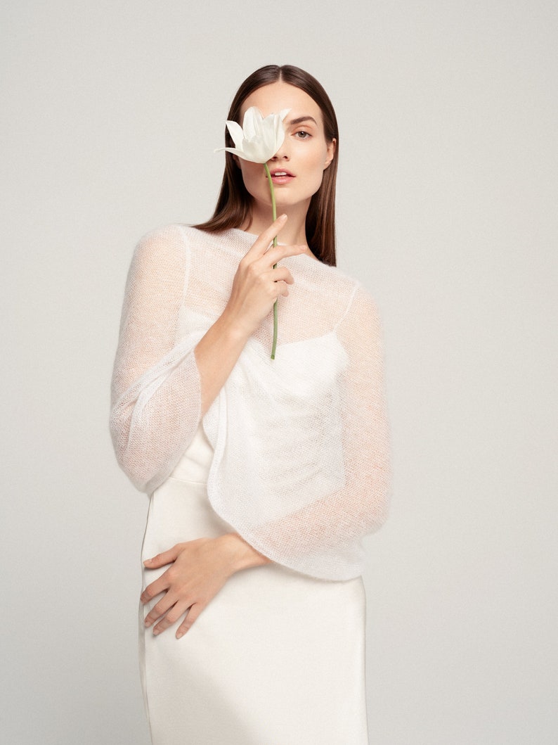 May include: A white sheer knit bolero with long sleeves and a wide neckline, worn over a solid white sleeveless top. The garment features a lightweight, semi-transparent texture and a clean, simple design that sits comfortably over the shoulders.