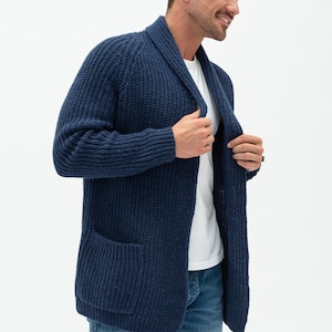 May include: A navy blue knitted cardigan with a shawl collar and a patch pocket on the left side. The cardigan is open and the person is wearing a white t-shirt and blue jeans underneath.