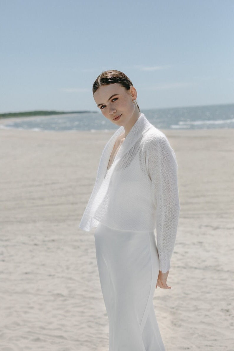 May include: A white knitted bolero cardigan with long sleeves and an open front, worn over a matching white sleeveless dress. The bolero features a textured, lightweight knit pattern and a shawl collar, providing a layered look.