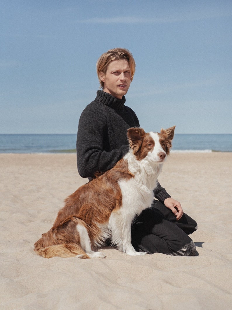 Puede incluir: Un hombre con un su&eacute;ter negro de cuello alto se sienta en una playa de arena con un perro border collie marr&oacute;n y blanco.