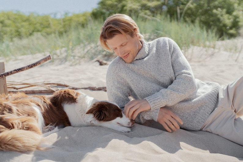 Puede incluir: Un hombre con un su&eacute;ter gris claro se sienta en una playa de arena con un perro marr&oacute;n y blanco. El hombre est&aacute; acariciando al perro, que est&aacute; acostado de lado.
