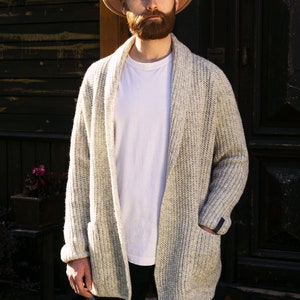 May include: A man wearing a light brown fedora hat, a white t-shirt, a light gray cardigan, and black pants. He is standing in front of a wooden door.