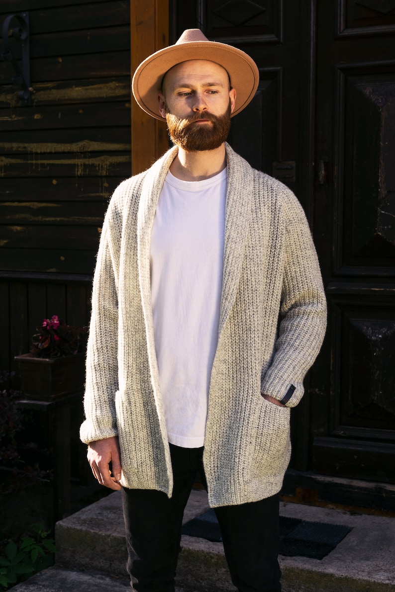 May include: A man wearing a light brown fedora hat, a white t-shirt, a light gray cardigan, and black pants. He is standing in front of a wooden door.
