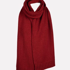 May include: A deep red knitted scarf featuring a consistent ribbed texture throughout the main body and finished with wide, vertical ribbed edges at both ends. The scarf is looped once at the top, creating a soft, folded drape that hangs vertically.