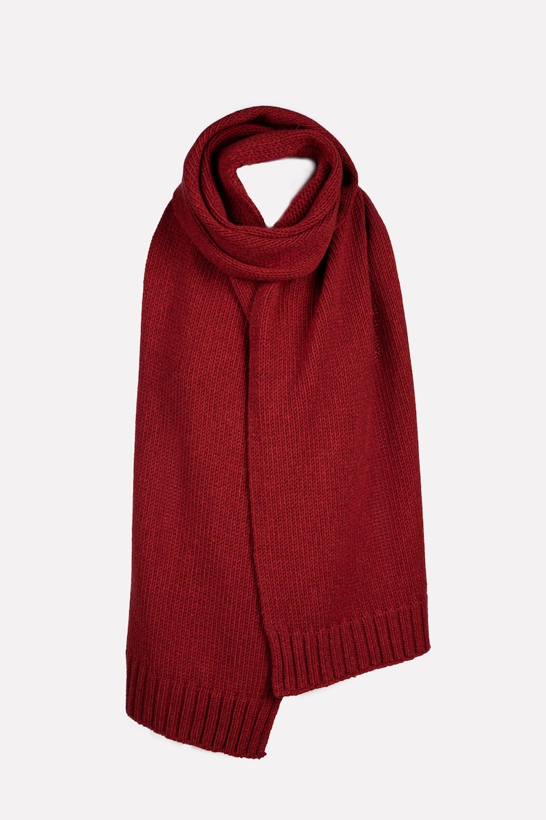 May include: A deep red knitted scarf featuring a consistent ribbed texture throughout the main body and finished with wide, vertical ribbed edges at both ends. The scarf is looped once at the top, creating a soft, folded drape that hangs vertically.