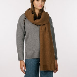 May include: A brown ribbed knit beanie and a matching long brown knit scarf with ribbed ends, worn over a grey long-sleeved sweater and blue denim jeans. The set features a consistent brown color and a soft, textured knit construction throughout.
