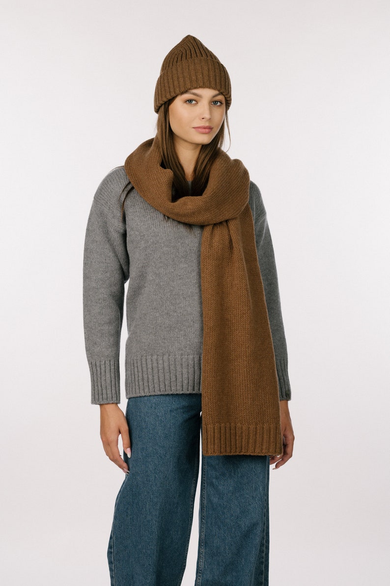 May include: A brown ribbed knit beanie and a matching long brown knit scarf with ribbed ends, worn over a grey long-sleeved sweater and blue denim jeans. The set features a consistent brown color and a soft, textured knit construction throughout.