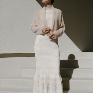 May include: A cream-colored, floor-length dress featuring a textured, smocked bodice and skirt with a ruffled hem, paired with a soft, long-sleeved, open-front beige cardigan and metallic gold strappy sandals with a square toe design.