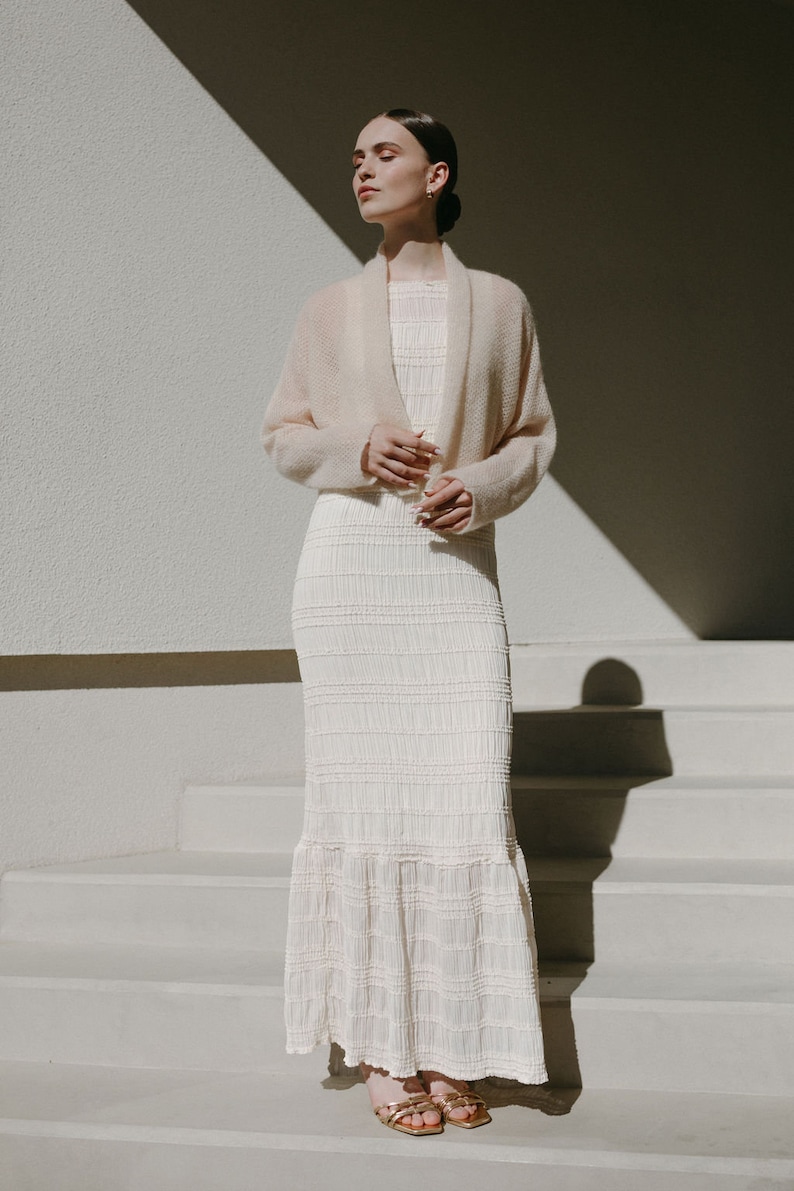 May include: A cream-colored, floor-length dress featuring a textured, smocked bodice and skirt with a ruffled hem, paired with a soft, long-sleeved, open-front beige cardigan and metallic gold strappy sandals with a square toe design.