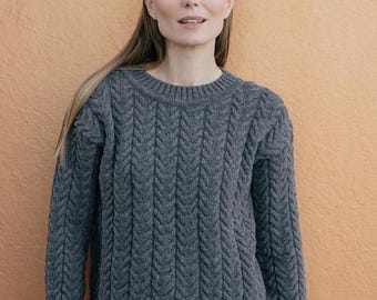 Puede incluir: Un suéter de manga larga en color gris carbón con un patrón de tejido de trenzas vertical, cuello redondo y puños y dobladillo acanalados. La prenda presenta un diseño de punto texturizado que recorre uniformemente el frente y las mangas.