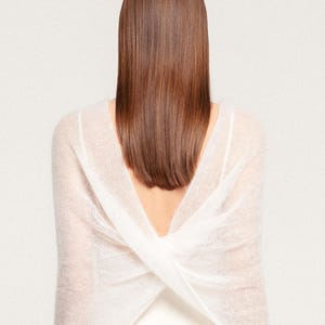 May include: A white, sheer, knitted wrap featuring a cross-over design at the back. The garment is constructed from a lightweight, semi-transparent material with a soft, fuzzy texture, creating a draped effect across the shoulders and upper back.