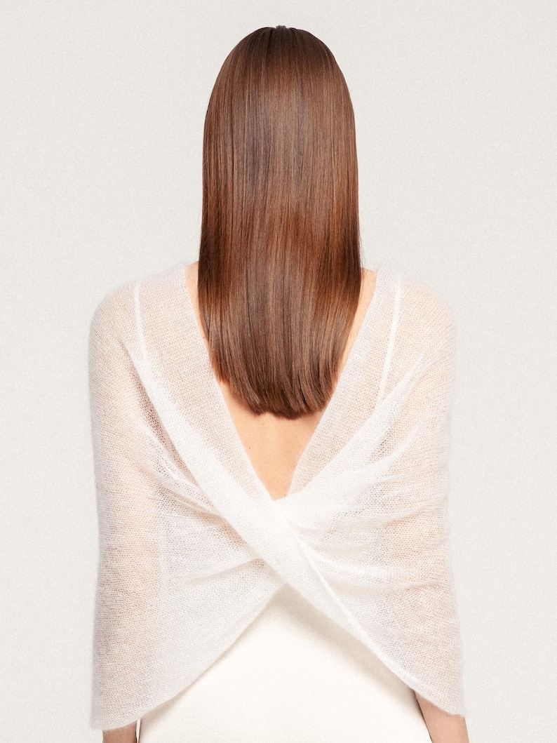 May include: A white, sheer, knitted wrap featuring a cross-over design at the back. The garment is constructed from a lightweight, semi-transparent material with a soft, fuzzy texture, creating a draped effect across the shoulders and upper back.