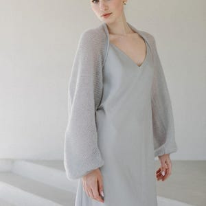 May include: A light gray slip dress with a V-neckline paired with a matching light gray bolero shrug. The shrug features long, voluminous sleeves made from a fine, open-knit, semi-sheer fabric that contrasts with the smooth, solid texture of the dress.