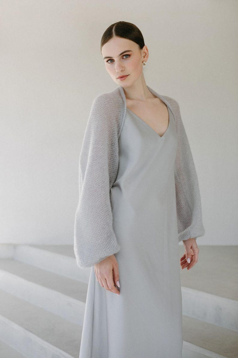 May include: A light gray slip dress with a V-neckline paired with a matching light gray bolero shrug. The shrug features long, voluminous sleeves made from a fine, open-knit, semi-sheer fabric that contrasts with the smooth, solid texture of the dress.