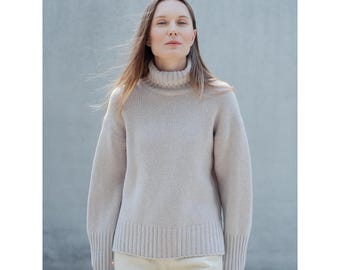 May include: A light beige turtleneck sweater with ribbed cuffs and hem. The sweater is made of a soft, knitted material. The model is wearing beige pants. The background is a light gray wall.
