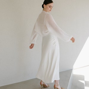 May include: A white midi-length skirt paired with a matching sheer, long-sleeved knit bolero top. The outfit is completed with gold-toned strappy high-heeled sandals, creating a coordinated ensemble with a clean, solid color palette.