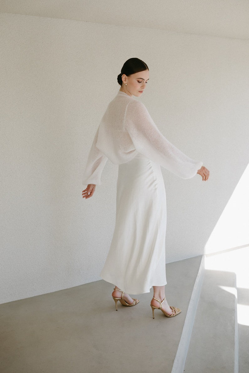 May include: A white midi-length skirt paired with a matching sheer, long-sleeved knit bolero top. The outfit is completed with gold-toned strappy high-heeled sandals, creating a coordinated ensemble with a clean, solid color palette.