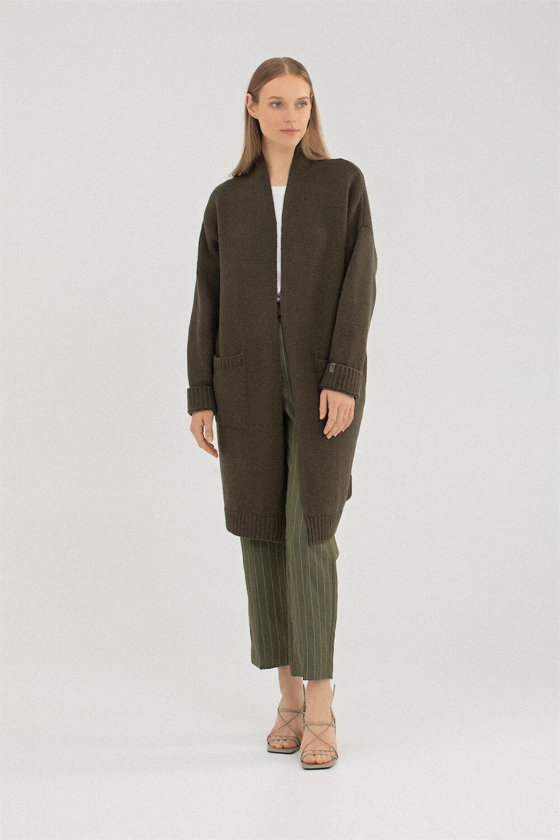 May include: A long, dark green cardigan with a shawl collar and pockets. The cardigan is made of a soft, knitted fabric.