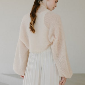 May include: A cream-colored, cropped knit bolero with long, voluminous sleeves and a textured, open-weave pattern, paired with a long, flowing, pleated skirt in a matching light cream shade, creating a soft and layered ensemble.