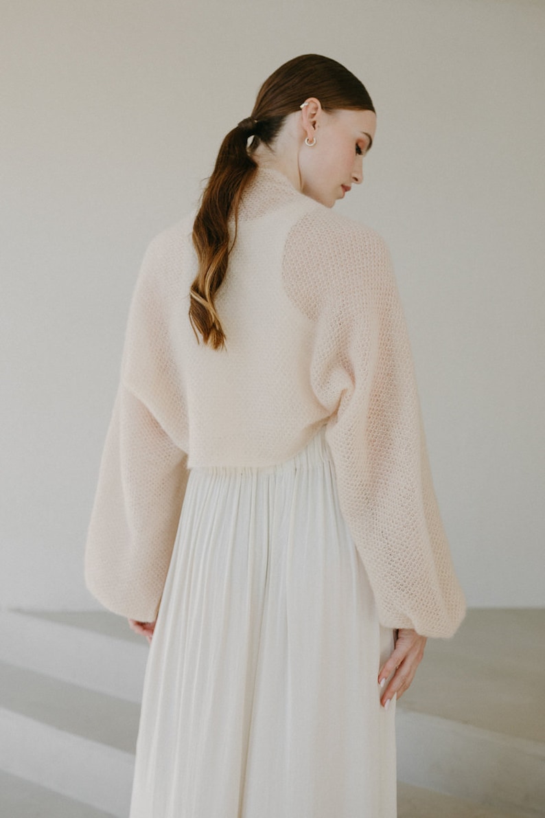May include: A cream-colored, cropped knit bolero with long, voluminous sleeves and a textured, open-weave pattern, paired with a long, flowing, pleated skirt in a matching light cream shade, creating a soft and layered ensemble.
