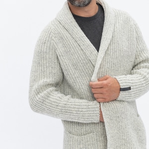 May include: A light gray knitted cardigan with a shawl collar and a single button closure. The cardigan has a patch pocket on the left side and a small black label on the right sleeve.