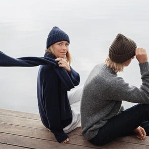May include: A navy blue ribbed knit beanie and matching scarf worn with a navy blue sweater and white trousers, alongside a brown ribbed knit beanie worn with a grey crewneck sweater and dark blue jeans, all featuring textured knit patterns.
