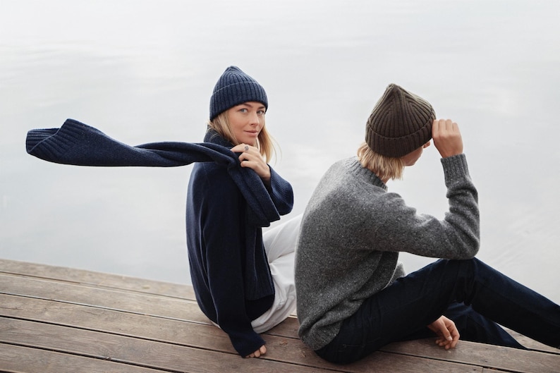 May include: A navy blue ribbed knit beanie and matching scarf worn with a navy blue sweater and white trousers, alongside a brown ribbed knit beanie worn with a grey crewneck sweater and dark blue jeans, all featuring textured knit patterns.