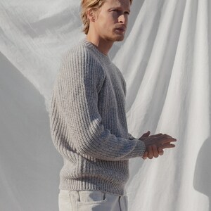May include: A man wearing a light gray knitted sweater with a crew neck and long sleeves. He is wearing white jeans and is standing in front of a white backdrop.