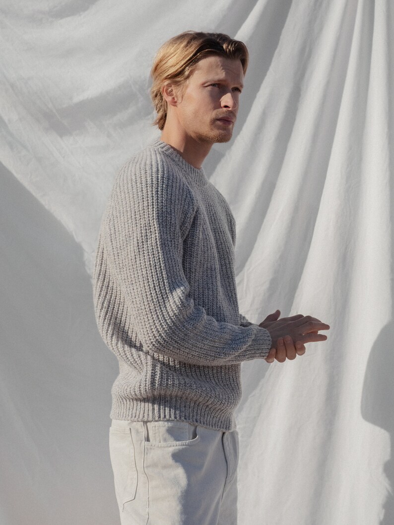 May include: A man wearing a light gray knitted sweater with a crew neck and long sleeves. He is wearing white jeans and is standing in front of a white backdrop.