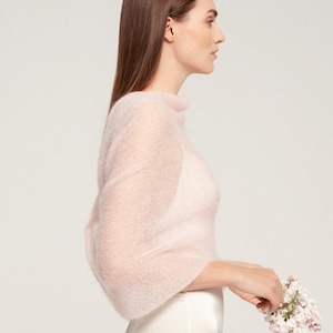May include: A light pink, sheer, long-sleeved shawl with a high neckline. The shawl is made of a soft, delicate material. The model is holding a small bouquet of white and pink flowers.