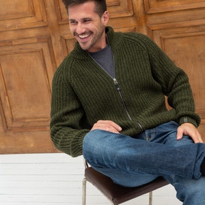 Pu&ograve; includere: Un uomo indossa un cardigan verde scuro a zip in maglia e jeans blu, seduto su una sedia marrone con struttura in metallo. Sorride e guarda a destra.