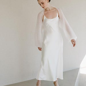 May include: A floor-length white satin slip dress with thin spaghetti straps and a scoop neckline, paired with a sheer, long-sleeved white knit bolero. The outfit is completed with gold-toned strappy high-heeled sandals with an open toe.