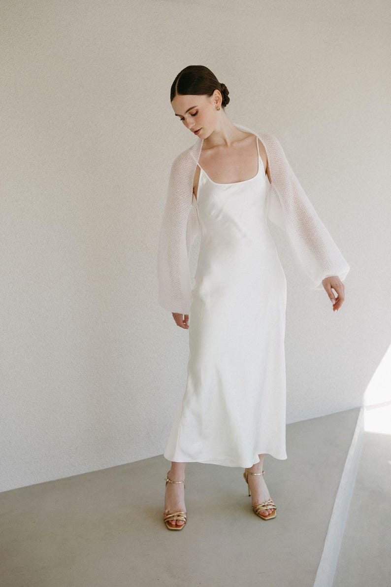 May include: A floor-length white satin slip dress with thin spaghetti straps and a scoop neckline, paired with a sheer, long-sleeved white knit bolero. The outfit is completed with gold-toned strappy high-heeled sandals with an open toe.