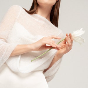 May include: A sheer white knitted sweater with a wide boat neckline and three-quarter length sleeves. The garment features a lightweight, semi-transparent texture and is worn over a solid white base layer, paired with a single white tulip flower.