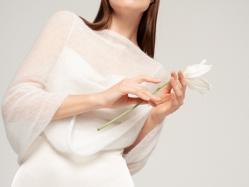 May include: A sheer white knitted sweater with a wide boat neckline and three-quarter length sleeves. The garment features a lightweight, semi-transparent texture and is worn over a solid white base layer, paired with a single white tulip flower.