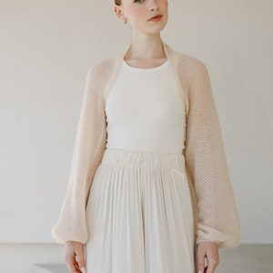 May include: A beige, sheer, open-front bolero shrug with long, voluminous sleeves and a fine knit texture, worn over a white ribbed tank top and a long, flowing white skirt with a gathered elastic waistband.