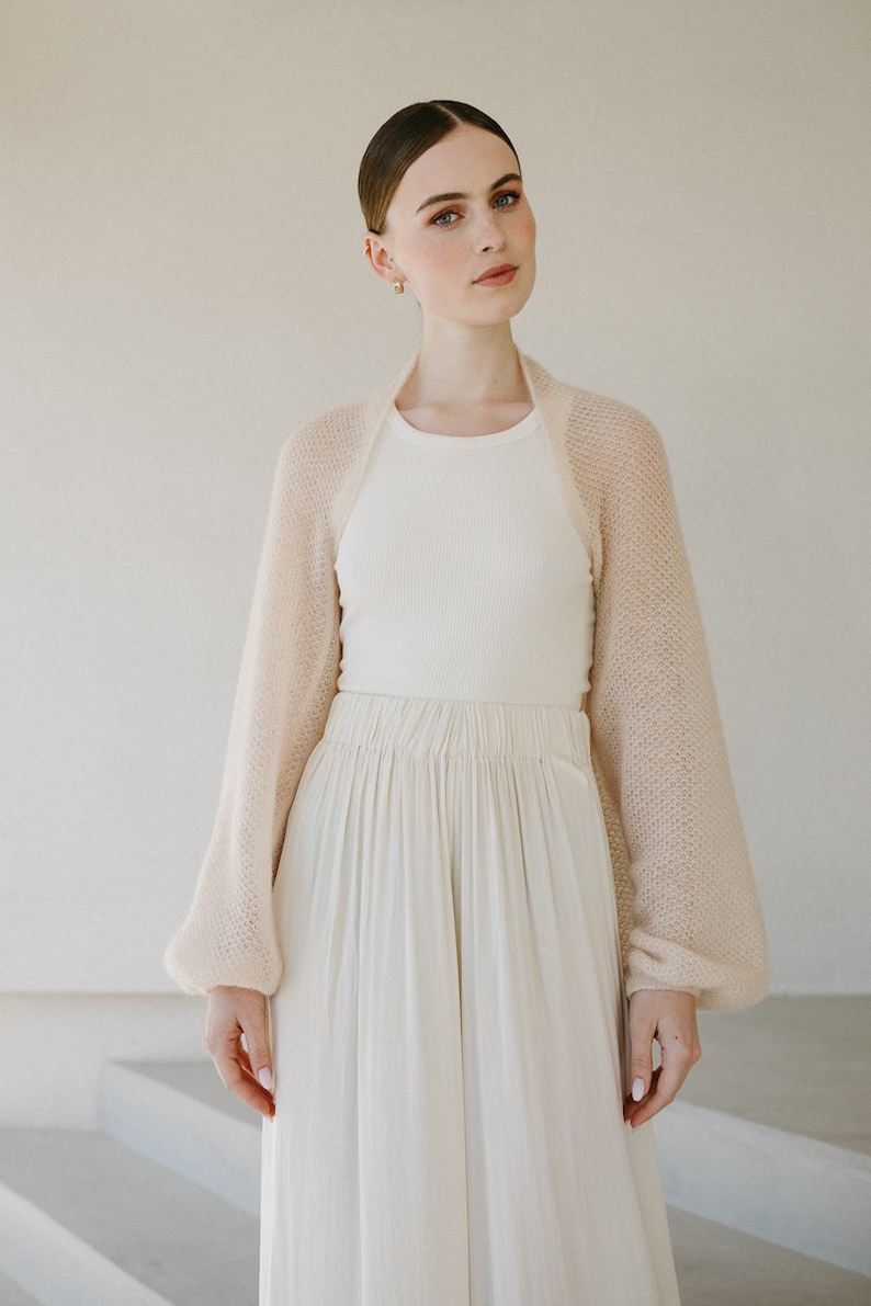 May include: A beige, sheer, open-front bolero shrug with long, voluminous sleeves and a fine knit texture, worn over a white ribbed tank top and a long, flowing white skirt with a gathered elastic waistband.
