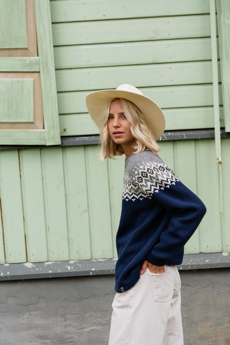May include: A woman wearing a blue and grey patterned sweater with a white hat. The sweater has a fair isle design. She is standing in front of a green wooden wall.