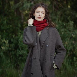 May include: A dark gray double-breasted wool coat with a notched lapel and dark buttons, worn over a light olive green shirt and a vibrant red ribbed knit neck warmer that sits snugly around the neck, providing a layered and textured appearance.