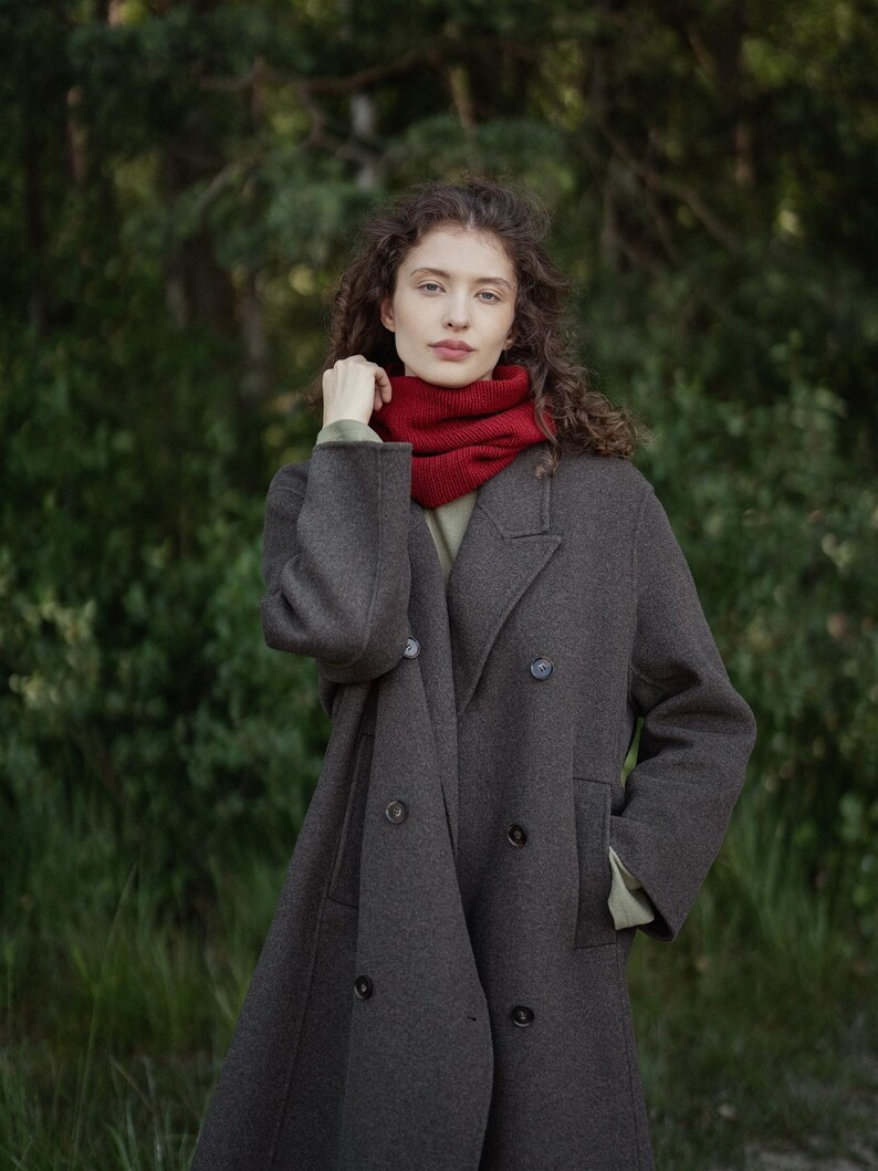 May include: A dark gray double-breasted wool coat with a notched lapel and dark buttons, worn over a light olive green shirt and a vibrant red ribbed knit neck warmer that sits snugly around the neck, providing a layered and textured appearance.