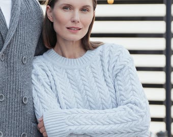 Puede incluir: Un suéter de punto trenzado azul claro con cuello redondo y puños acanalados, usado junto a un cárdigan gris de doble botonadura con cuello de chal y botones grandes. Ambas prendas están hechas de tejido grueso y texturizado.