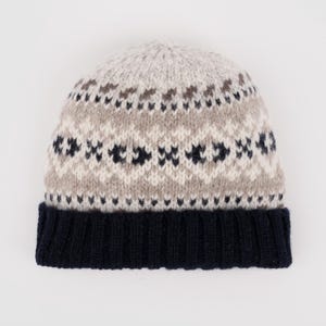 May include: A knitted beanie hat with a navy blue brim and a patterned crown. The crown features a repeating geometric design in light gray, brown, and navy blue. The hat is made of a soft, warm material.