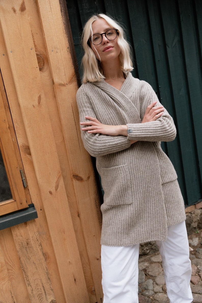 Pu&ograve; includere: Una donna indossa un cardigan di maglia marrone chiaro con cappuccio e pantaloni bianchi. &Egrave; in piedi accanto a un muro di legno e ha le braccia incrociate.