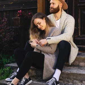 May include: A couple sitting on a set of stone steps outside a wooden building. The woman is wearing a beige cardigan and black leggings, while the man is wearing a beige cardigan over a white shirt and black pants. The man is wearing a brown fedora hat.