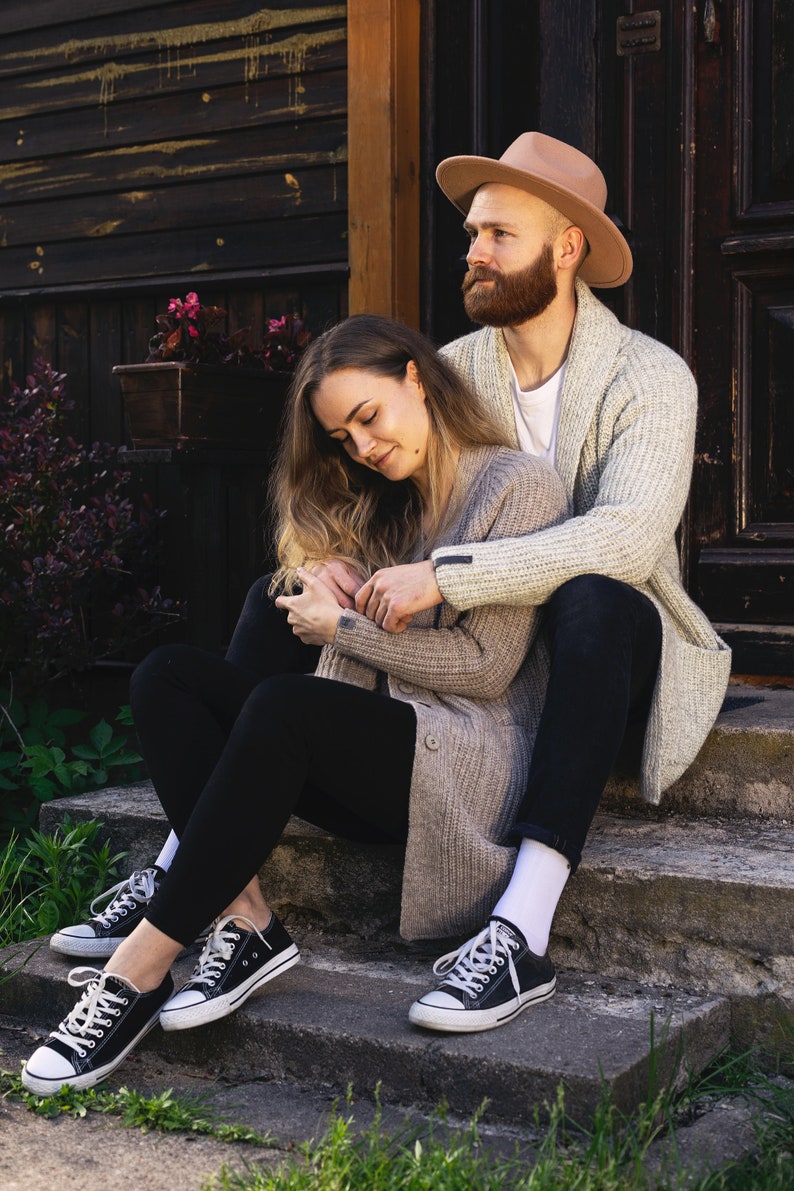 May include: A couple sitting on a set of stone steps outside a wooden building. The woman is wearing a beige cardigan and black leggings, while the man is wearing a beige cardigan over a white shirt and black pants. The man is wearing a brown fedora hat.