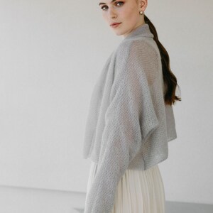 May include: A light gray, sheer, long-sleeved knit bolero with a textured, open-weave pattern and a high collar. The garment is cropped at the waist and features a relaxed fit with dropped shoulders, worn over a cream-colored, pleated skirt.
