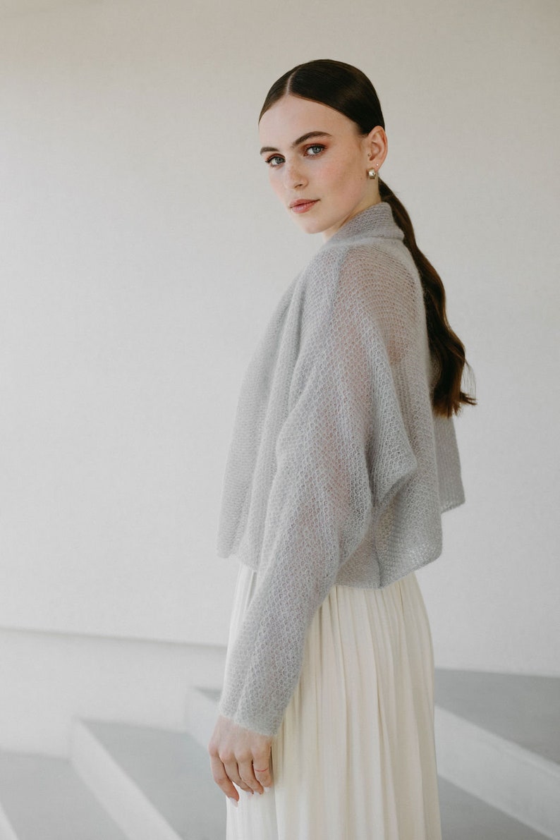 May include: A light gray, sheer, long-sleeved knit bolero with a textured, open-weave pattern and a high collar. The garment is cropped at the waist and features a relaxed fit with dropped shoulders, worn over a cream-colored, pleated skirt.