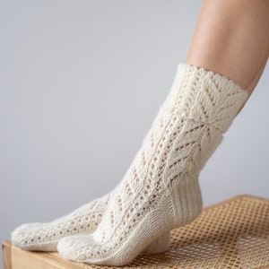 May include: A pair of white knitted socks with a lacy pattern. The socks are on a wooden stool with a woven seat.